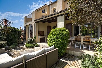 A sunny patio of a Mediterranean-style house with outdoor seating, dining table, and lush greenery under a bright blue sky.