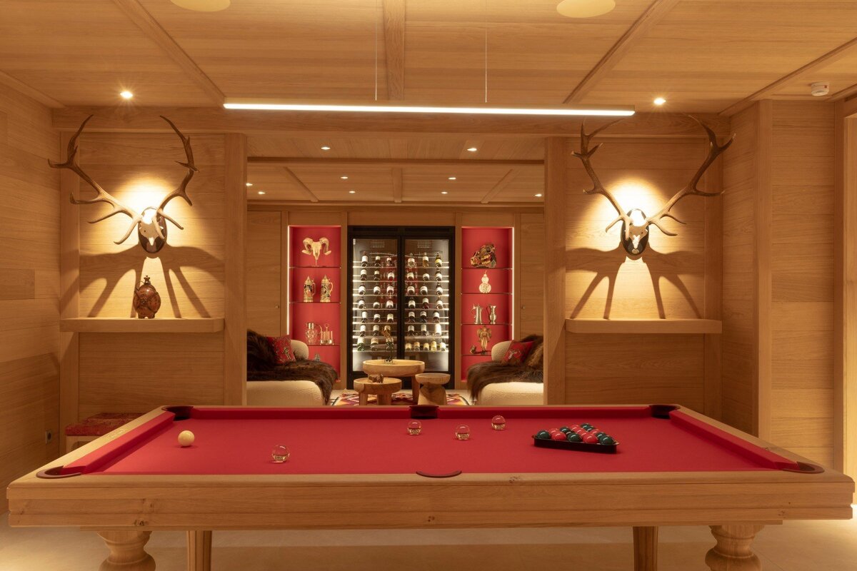 A chic wooden room with a red billiard table, antler wall decor, and a lounge with a large wine cooler and red display cases.