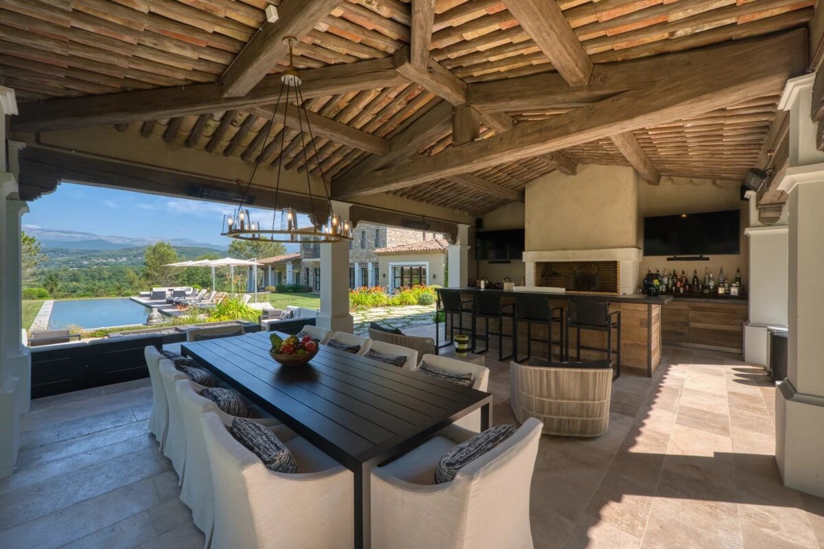 Luxurious covered patio with a long dining table, full bar, fireplace, and an infinity pool, all set against a scenic mountain view.