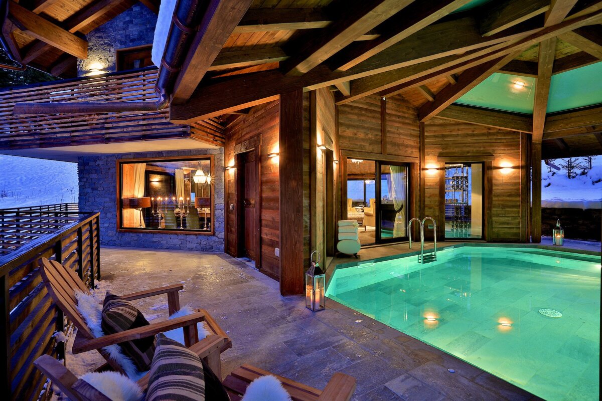 Luxurious wooden chalet with an inviting turquoise pool, cozy seating, and warm lighting. Snowy mountains provide a serene winter backdrop.