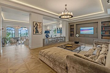 Elegant living room with a large sectional sofa, built-in TV unit & bookshelves, and a chandelier. Arched windows overlook mountains and sea.