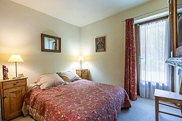 A cozy bedroom with a floral bedspread, two nightstands with lamps, a mirror, and a glass door with red floral curtains beside a wooden chair.