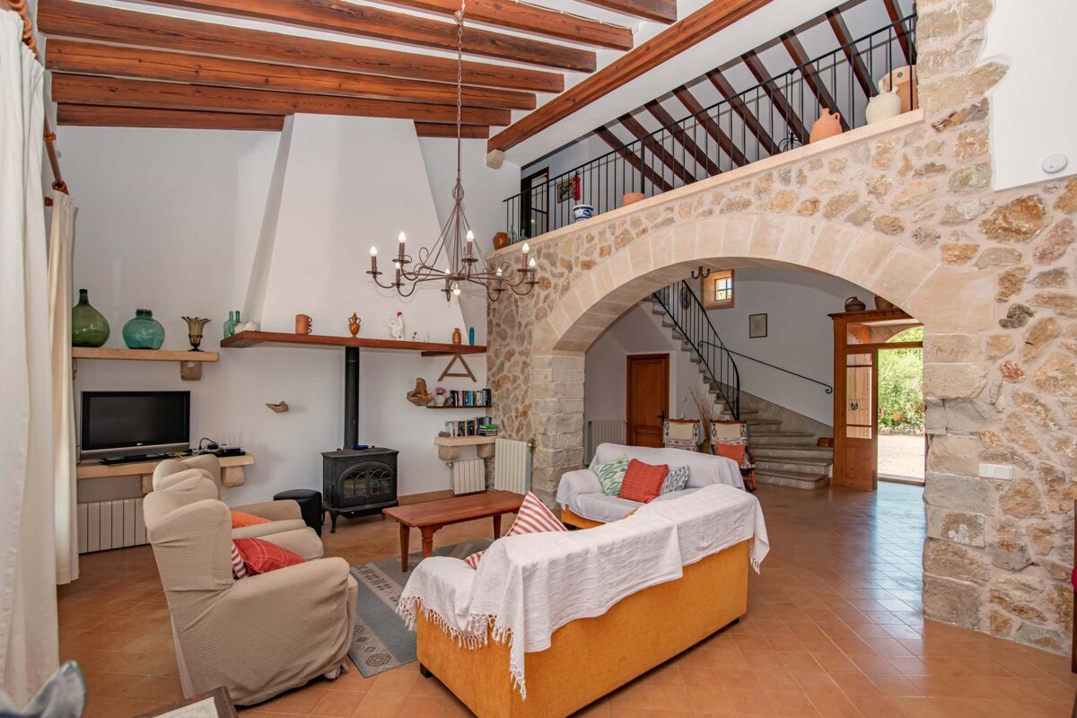 A rustic living room with stone walls, wooden beams, fireplace, and arched entryway leading to a staircase and mezzanine. Sofas fill the cozy space.