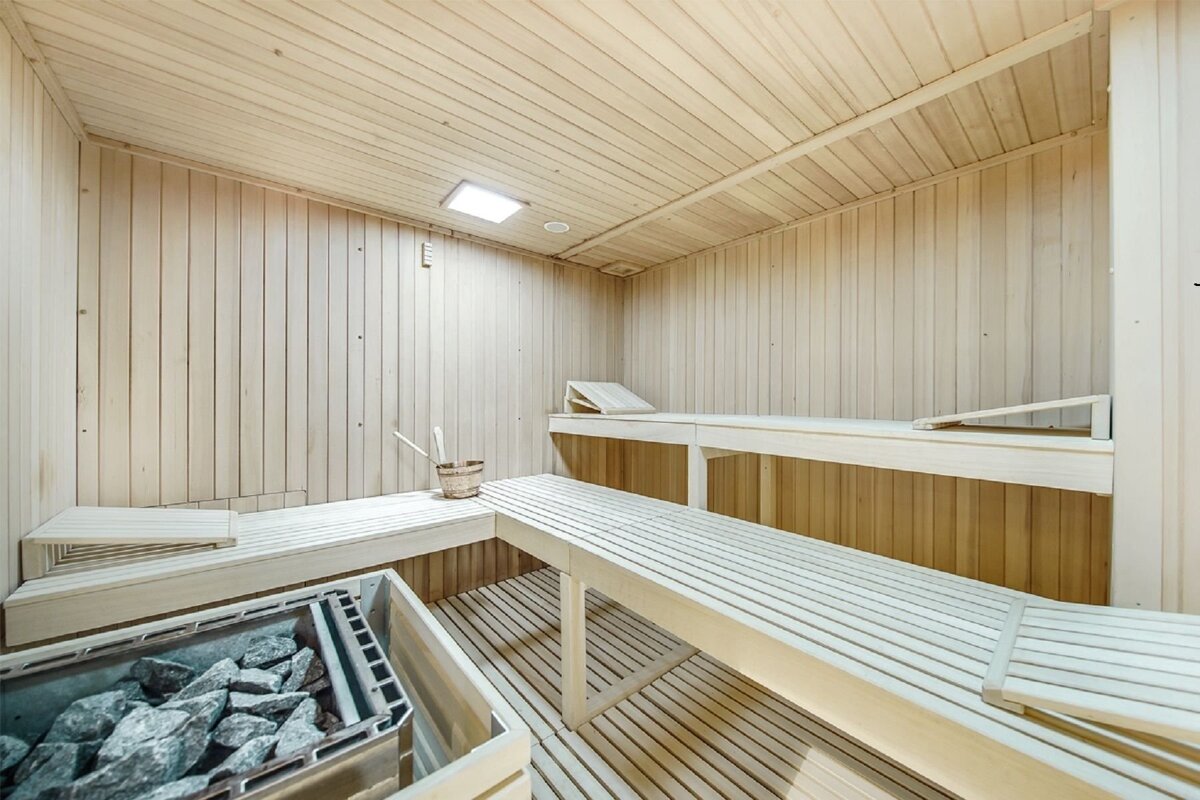 A pristine, light wood sauna features multiple benches, a rock heater, and a traditional bucket and ladle, with a bright light on the ceiling.