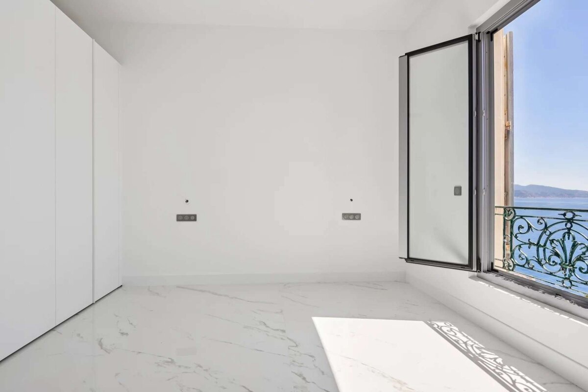 Bright, empty white room with marble floor & built-in closet. Open window shows a beautiful sea/mountain view & ornate balcony railing.