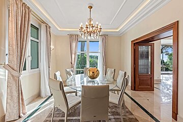 Luxurious dining room with a round glass table, white chairs, and a grand crystal chandelier. Cream curtains frame large windows, marble floor.