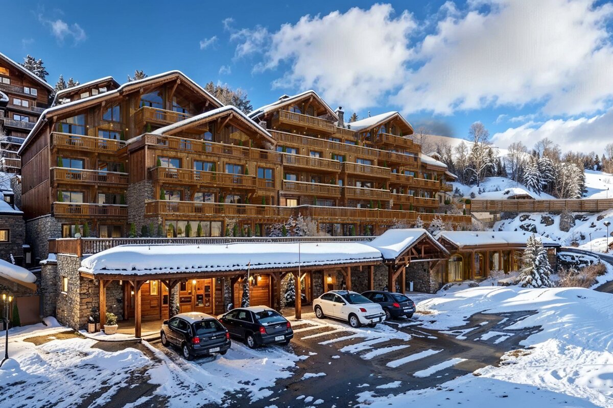 A large, luxurious wooden chalet hotel covered in snow, set in a winter mountain landscape under a blue sky. Cars are parked in front.
