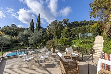 A sunny outdoor patio with ornate white and wicker furniture, overlooking a small pool and a lush green hillside under a blue sky.