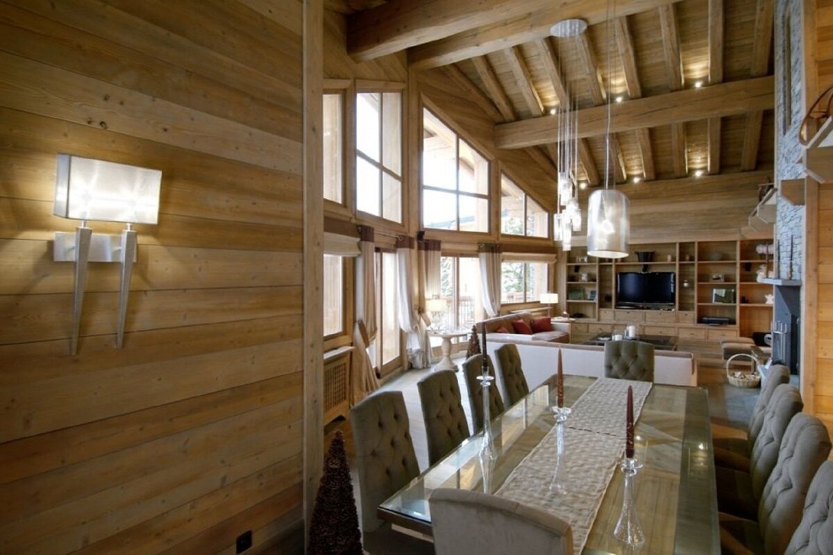 A large, rustic yet modern chalet interior featuring warm wood walls/ceilings, a long dining table, cozy living space, and expansive windows.