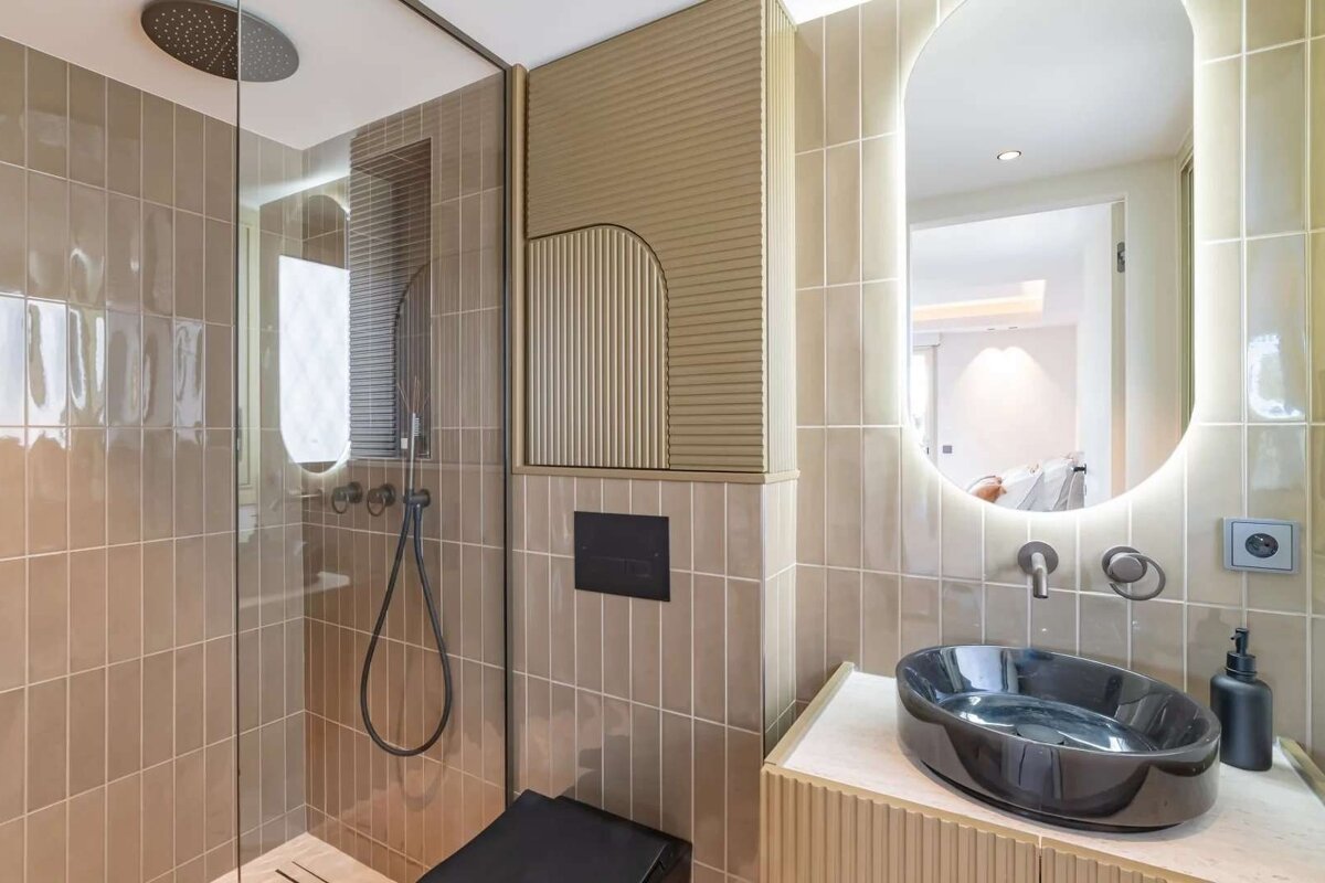 Modern beige-tiled bathroom with a glass shower, black oval sink, and an illuminated oval mirror. Stylish fluted paneling adds texture.
