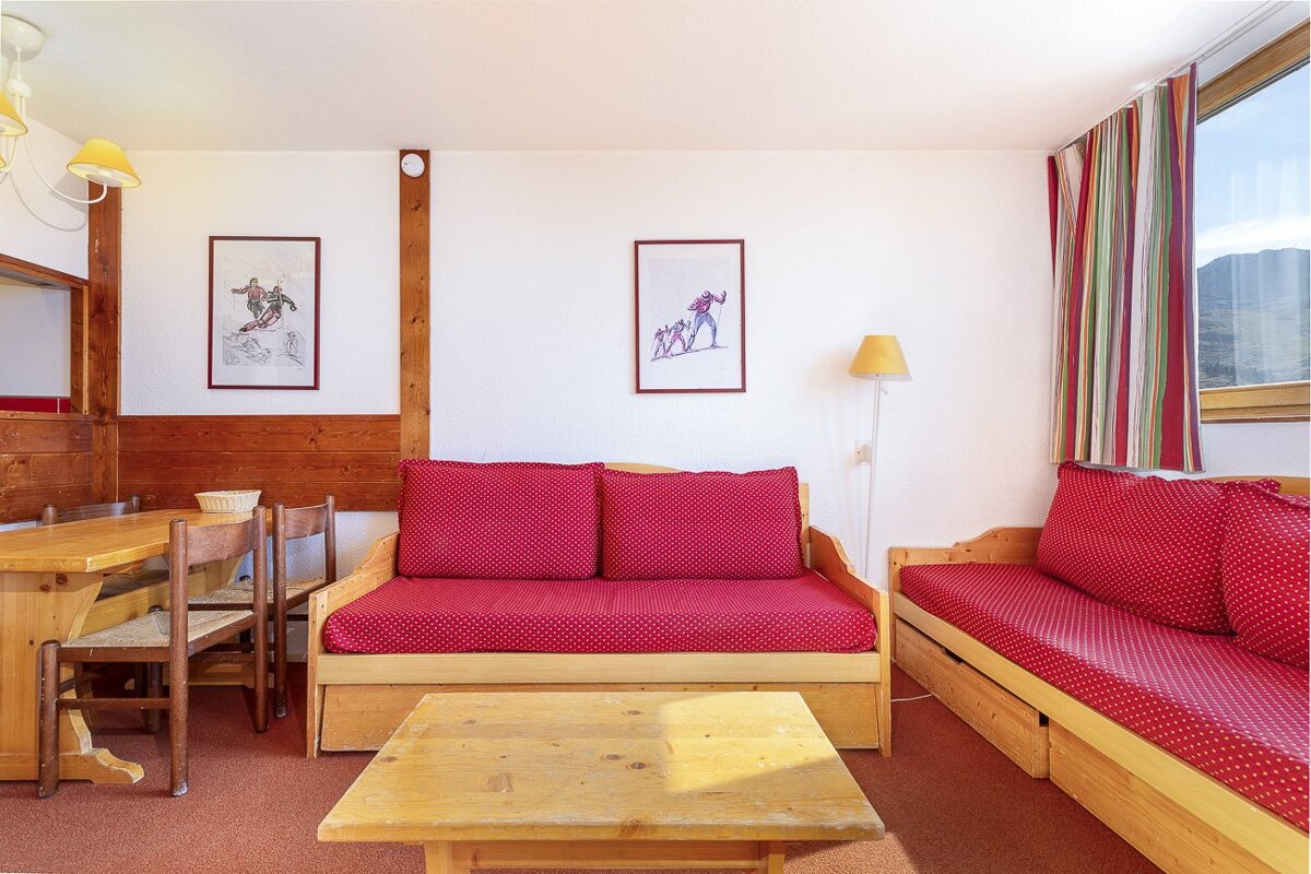 Cozy, rustic living room with wooden furniture, red polka dot sofas, ski-themed art, and a dining area, featuring a window with mountain views.