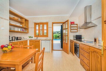 A bright kitchen featuring wooden cabinets, a dining table with a fruit bowl, and an open door leading to an outdoor area with a pool.