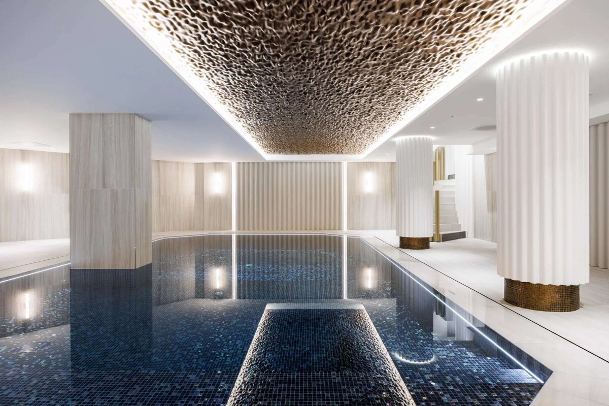 A luxurious indoor swimming pool features a textured gold ceiling, soft lighting, and elegant white fluted columns, creating a serene, upscale ambiance.