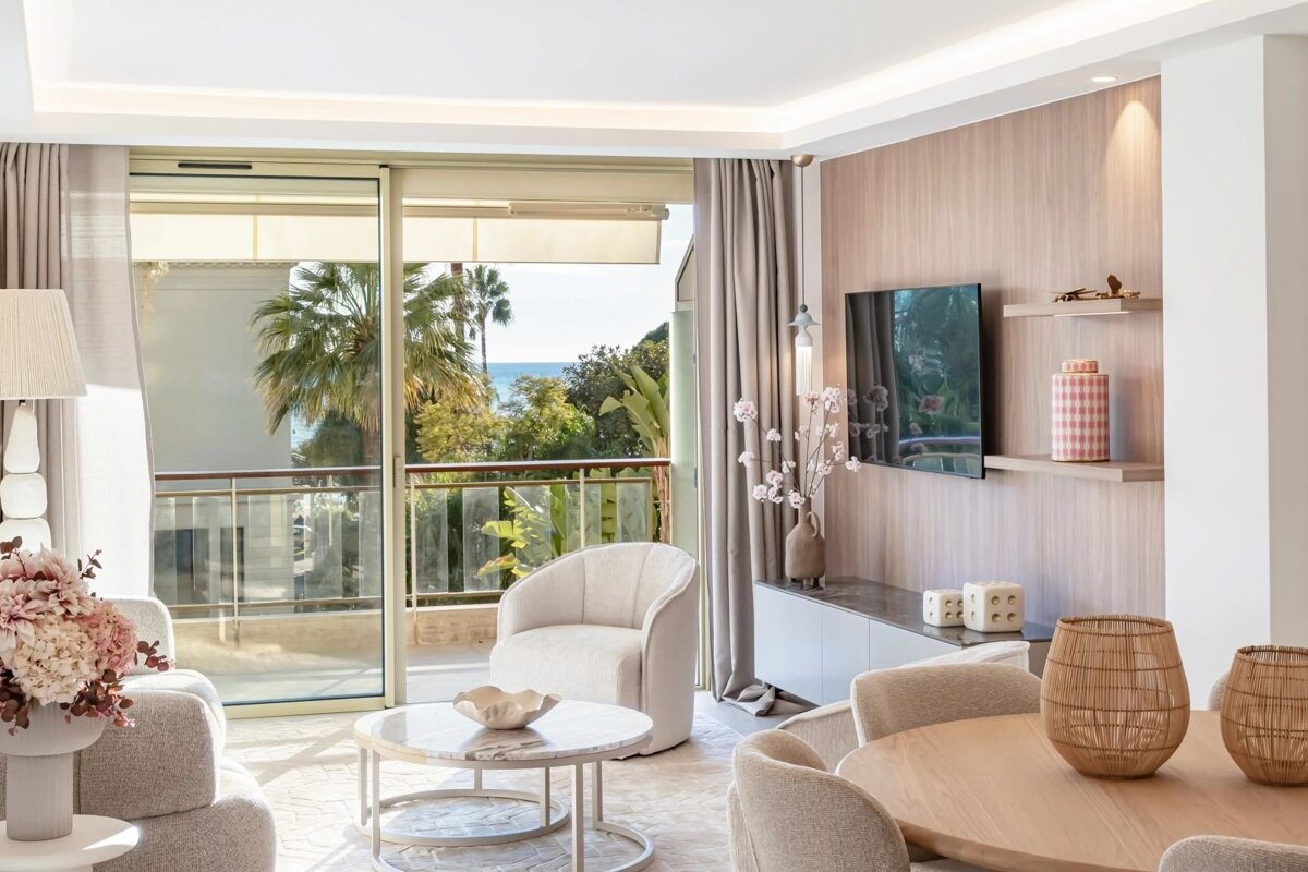 A bright, chic room with a balcony overlooking palm trees and the sea. Neutral decor, modern furniture, and a wall-mounted TV.