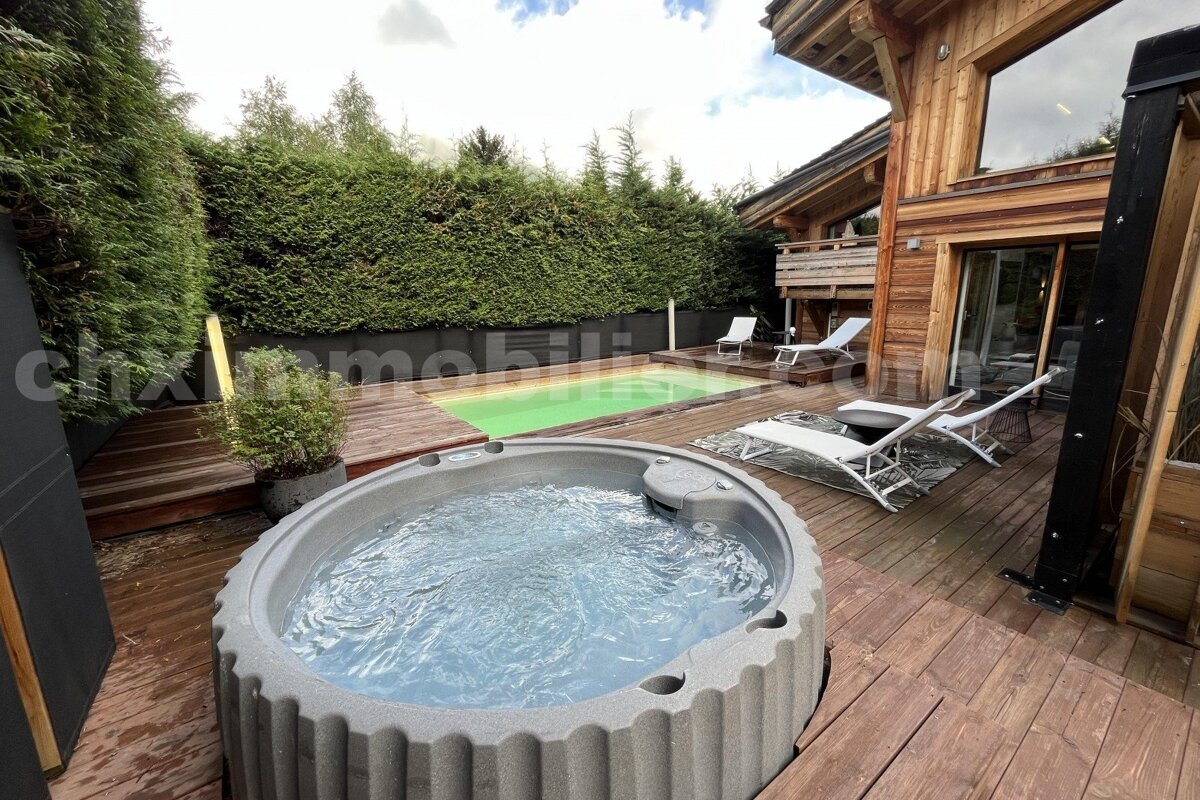 A luxurious outdoor space featuring a bubbling hot tub, small green pool, and wooden deck beside a modern wooden chalet, surrounded by hedges.