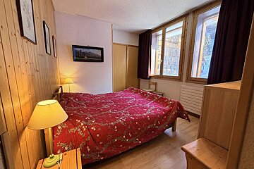 A cozy bedroom with a red floral bedspread, wood paneling, and two windows. Lamps and wall art complete the rustic look.