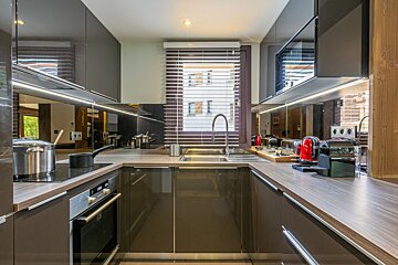 A sleek, modern U-shaped kitchen features glossy grey cabinets, wooden countertops, a window with blinds, reflective backsplash, and red appliances.