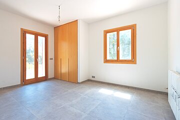 A bright, empty room with white walls, gray tiled floor, a wooden built-in wardrobe, and wooden-framed balcony doors and window. Sunlight falls on the floor.
