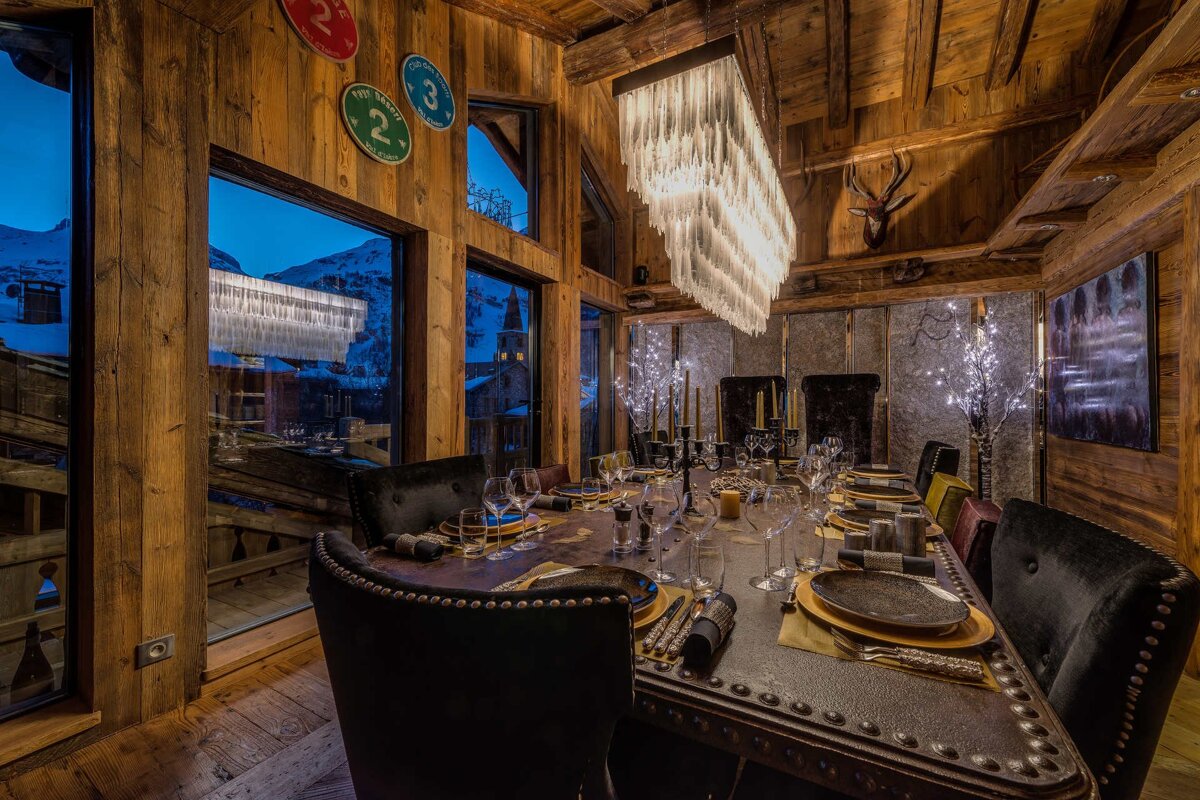 Cozy dining room in a rustic wooden chalet with a large icicle chandelier and window views of snowy mountains at dusk.