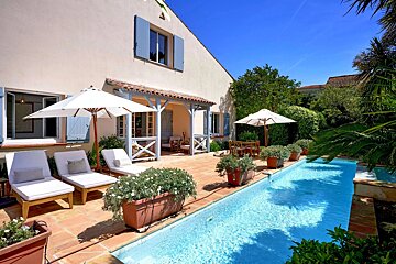 A Mediterranean-style house with blue shutters, a sun-drenched terracotta patio, loungers, and a long swimming pool, surrounded by lush plants.