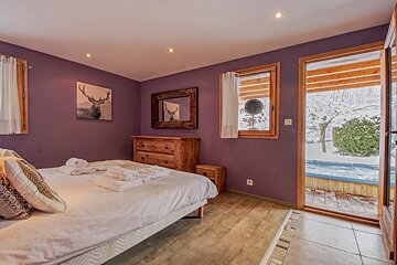 Cozy purple bedroom with a bed, wooden furniture, and deer art, opening to a snowy patio with an outdoor hot tub.