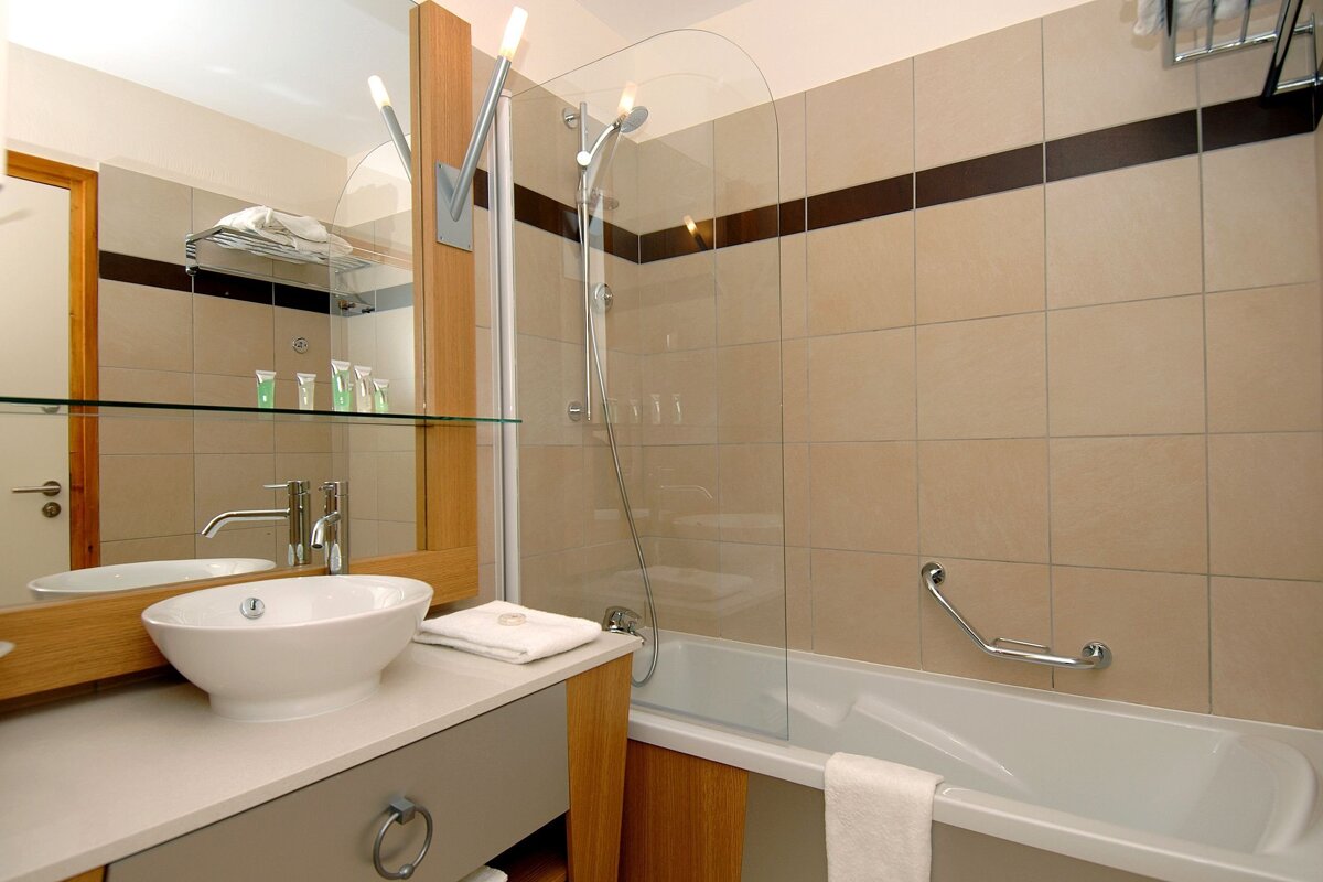 A modern bathroom with beige tiled walls, a dark accent stripe, a white vessel sink on a wooden vanity, and a bathtub with a glass shower screen.