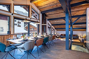 Modern alpine chalet dining area with a long table, colorful chairs, and large windows showing a snowy mountain landscape.