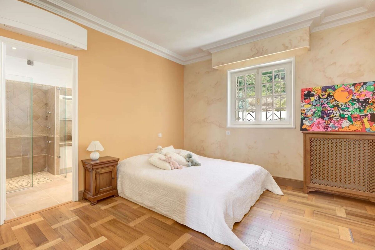 A peach-walled bedroom with a white bed, wood floor, colorful art, window, and an adjoining bathroom with a glass shower.