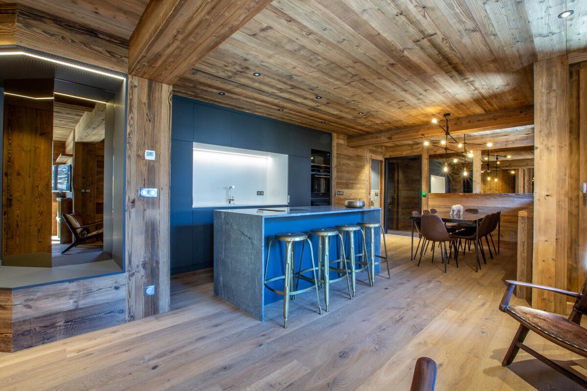 A modern chalet kitchen and dining area featuring warm wood paneling, a blue island with gold stools, and a dining table.