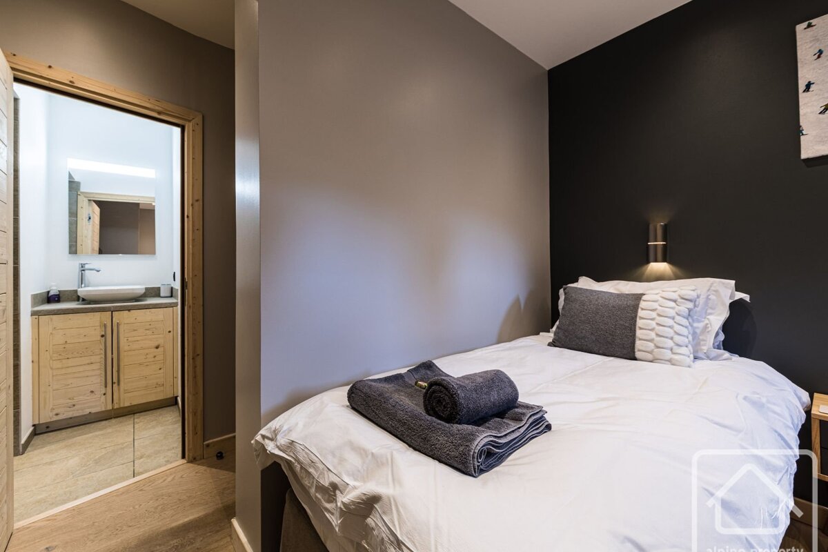 A cozy single bedroom with white bedding, grey towels, and a dark accent wall. An adjacent bathroom with a wooden vanity and sink is visible.