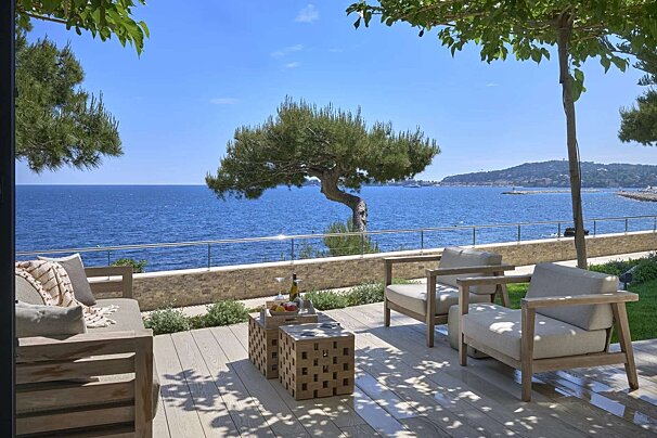 A sun-dappled patio with modern outdoor furniture and a table with refreshments overlooks a vibrant blue ocean, framed by a lone tree and distant land.