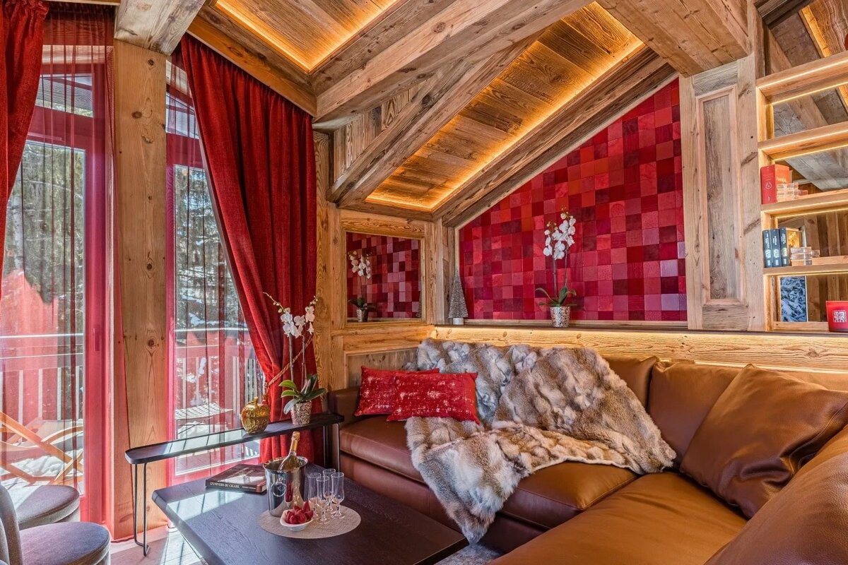 Cozy wooden chalet interior featuring a leather sofa, plush fur throw, warm lighting, and vibrant red accents like curtains and a tiled wall.