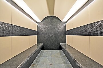 A modern steam room with black and beige mosaic tiles, built-in benches, strip lighting, and an arched wall.