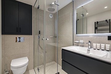 A modern, sleek bathroom with a glass shower, wall-mounted toilet, dark vanity with sink, and illuminated mirror against light-colored tiles.