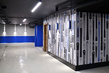 Stylish underground corridor with concrete floor, blue/grey walls, and a feature wall covered in ski-themed wallpaper. Modern lighting illuminates the space.