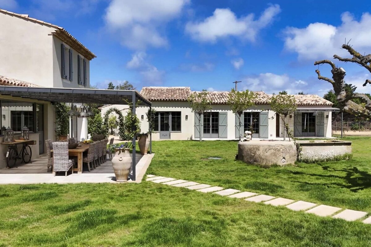 Sunny Mediterranean estate featuring an outdoor dining patio, lush green lawn, stepping stones, and charming white buildings with terracotta roofs.