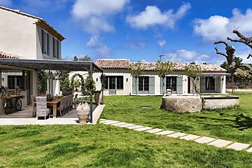 Sunny Mediterranean home with a large patio, outdoor dining, green lawn, stone well, and stepping path under a blue, cloudy sky.