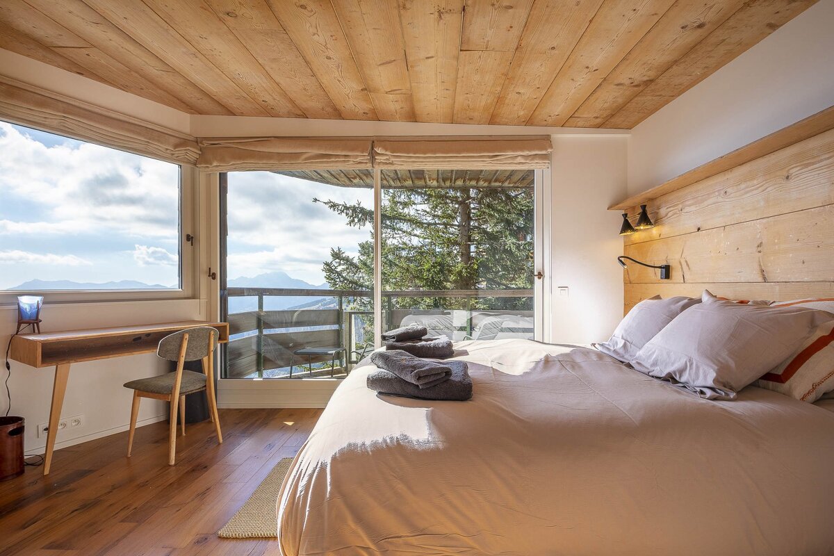 A bright bedroom with a wooden ceiling and floor, featuring large windows offering panoramic views of mountains and forests. A bed and desk complete the rustic-modern space.