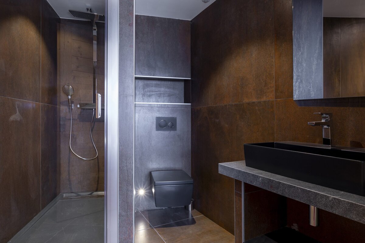 Modern bathroom with dark rustic tiled walls, a glass shower, wall-mounted toilet, and a black sink on a stone counter.