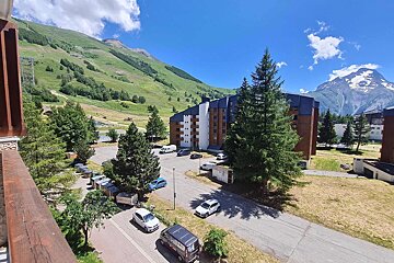 A sunny summer day at a mountain resort, with buildings, green slopes, pine trees, cars, and snow-capped peaks under a blue sky.