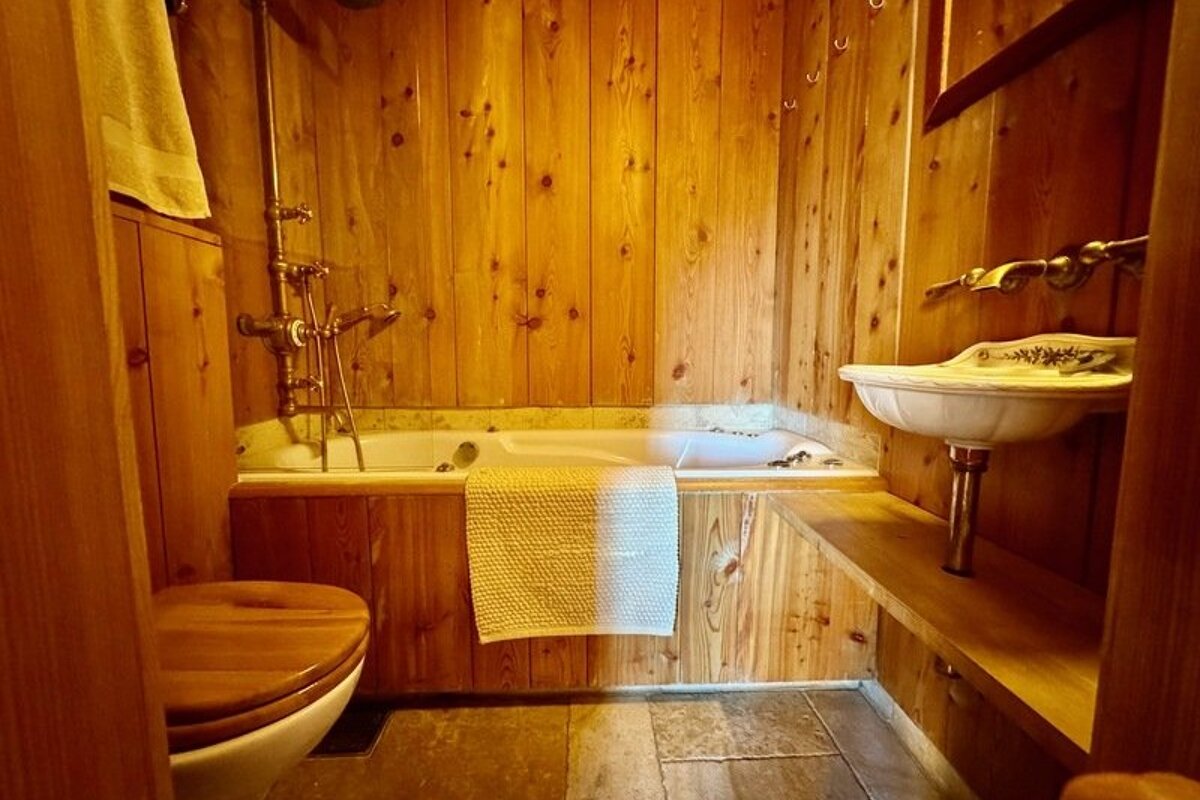 A cozy cabin bathroom featuring warm wood-paneled walls, a bathtub, a white sink, and a toilet with a wooden lid.