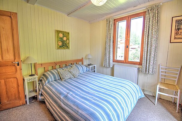 A cozy bedroom with yellow wood-paneled walls, a blue and white striped bed, a wooden-framed window with floral curtains, and a wooden door.