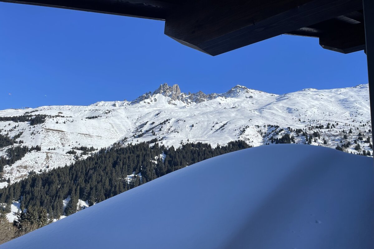 Snowy mountain vista with pine forests under a clear blue sky, viewed from a balcony with a prominent snowdrift in front.