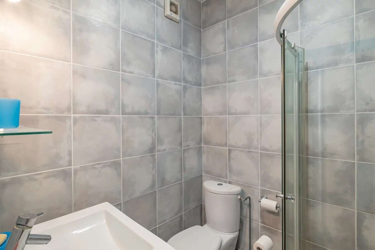 A modern bathroom with grey tiled walls, featuring a white sink, toilet, and glass shower enclosure. A blue cup sits on a glass shelf.