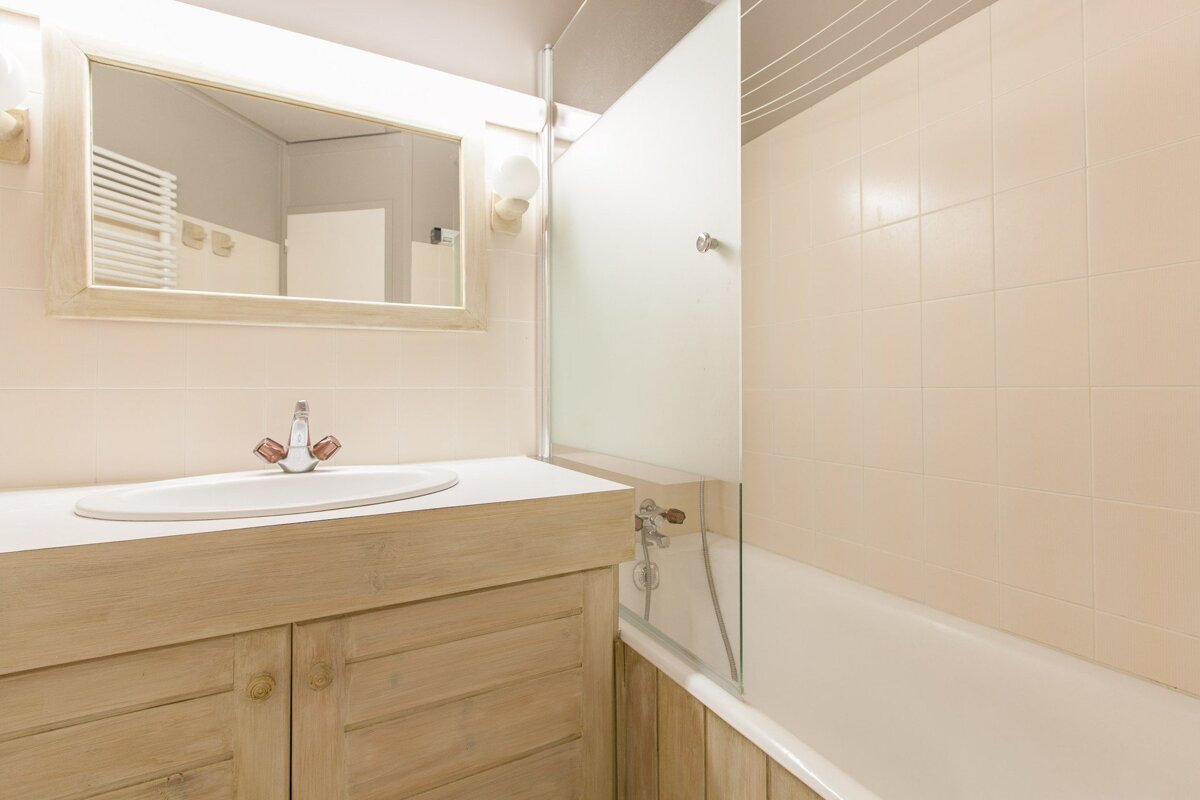 A bright bathroom with a light wood vanity, sink, mirror, and bathtub with a glass screen. Light tiled walls complete the look.