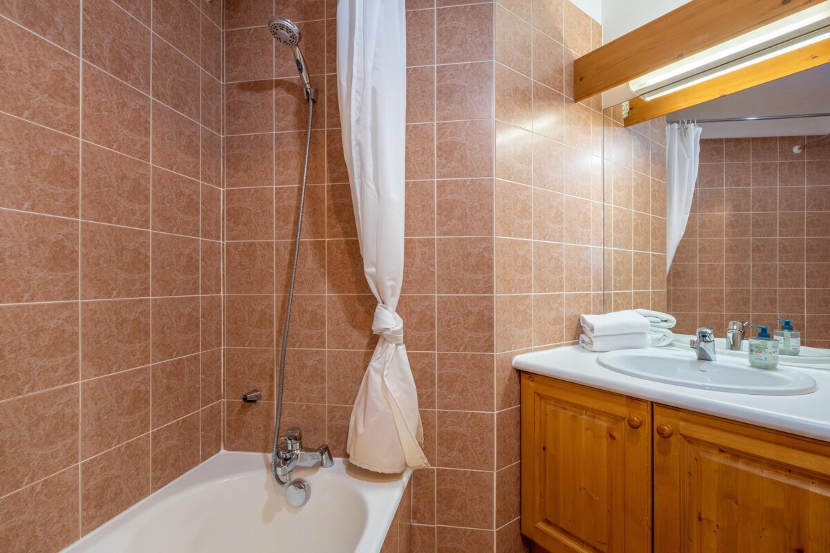 A functional bathroom with brown tiled walls, a bathtub/shower, a white curtain, and a vanity with a sink, mirror, and wooden cabinet.
