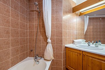 A functional bathroom with brown tiled walls, a bathtub/shower, a white curtain, and a vanity with a sink, mirror, and wooden cabinet.