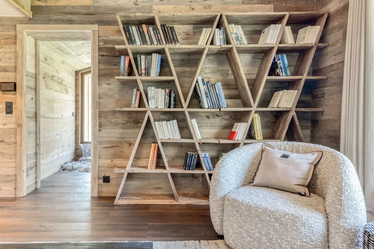 A cozy reading nook features a unique wooden geometric bookshelf and a textured boucle armchair with a pillow, set against rustic wood walls.
