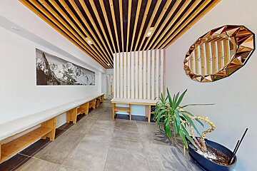 A modern hallway features a slatted wooden ceiling, a mountain mural, long wooden benches, a geometric mirror, and a potted plant on a tiled floor.