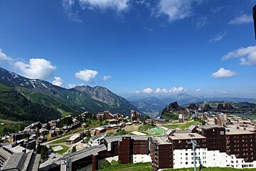 Panoramic shot of a mountain village with many buildings on green slopes, distant peaks with snow, and a vibrant blue sky with scattered clouds.